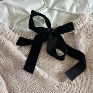 Ljusbeige stickad tröja med rosett - Superfin ljusbeige stickad tröja med rund hals och en svart sammetsrosett i nacken. Tröjan har en mysig, lite lös passform och långa ärmar. Perfekt för dig som gillar söta detaljer och vill ha något både stilrent och gulligt.