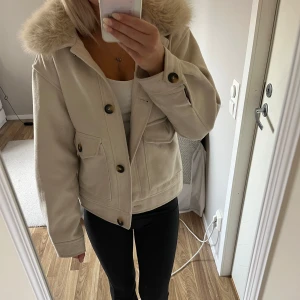 Beige jacka med päls och knappar - Snygg beige jacka med päls på kragen och stora dekorativa knappar. Jackan har två fickor framtill och är i en kortare modell med lång ärm. Perfekt för dig som vill ha en stilren och trendig look under höst och vinter. 