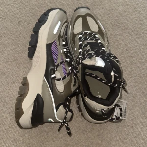 Chunky sneakers från H&M i grått och svart (oanvänd) - Säljer ett par chunky sneakers från H&M i grått, svart och beige med coola lila mesh-detaljer. Skorna har grov sula, svartvita snören och reflekterande inslag. Perfekta för dig som gillar streetstyle och vill sticka ut lite extra.