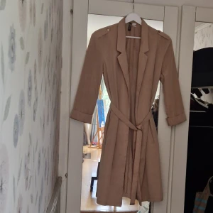Beige omlottskappa med bälte H&M - Stilren beige tunn kappa från H&M med knytbälte i midjan och trekvartsärmar. Kappan har klassisk krage. Perfekt för dig som gillar en enkel men trendig look. Passar mycket bra för kallare sommardagar eller sommarkväll. 