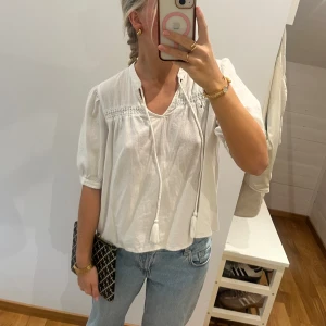 Vit blus med knyt och puffärm - Säljer en luftig vit blus med korta puffärmar och dekorativ knytning med tofsar framtill. Blusen har v-ringning och fina broderade detaljer vid axlarna. Perfekt till jeans för en chill och stilren look.