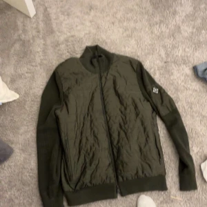 J lindberg cardigan  - Snygg olivgrön j lindberg cardigan  med quiltad framsida och stickade ärmar. Jackan har dragkedja hela vägen, ribbade muddar och en liten ficka med patch på ena ärmen. Perfekt för dig som vill ha en stilren och bekväm jacka till vardags.