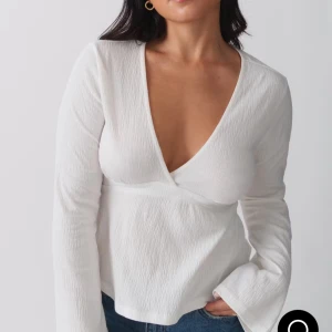 Vit omlottopp med knyt och v-ringning Gina - Säljer en vit omlottopp med djup v-ringning och långa ärmar. Toppen har en snygg knytning i ryggen och är gjord i ett lätt, strukturerat tyg som ger en stilren look. Perfekt att matcha med jeans för en fräsch och trendig outfit.