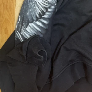 Marcelo Burlon (Äkta) - Svart sweatshirt med grått och vitt vingeprint över axlar och bröst. Tröjan har rund halsringning och långa ärmar. Mjuk och skön bomullskänsla, perfekt för dig som gillar streetstyle och unika detaljer.