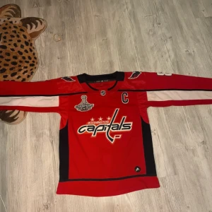 Washington Capitals Ovechkin #8 tröja - Röd långärmad hockeytröja från Adidas med Washington Capitals tryck, Ovechkin #8 på ryggen och Stanley Cup Champions-märke. Tröjan har svarta och vita detaljer samt broderade loggor på axlarna. Perfekt för dig som älskar hockey och vill sticka ut på läktaren.