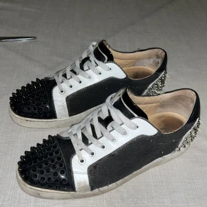 Christian Louboutin sneakers med nitar - Svarta och vita sneakers från Christian Louboutin med röda sulor, svarta nitar på tån och silvriga nitar på hälen. Skorna har snörning, rund tå och är tillverkade i skinn och mocka. Riktigt edgy vibe och sticker ut med sina detaljer. Dom är äkta men har ej låda pris kan diskuteras
