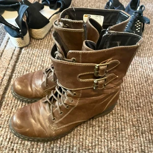 Bruna boots med spännen och snörning - Snygga bruna boots i skinn med snörning framtill och två dekorativa spännen på skaftet. Grov sula och dragkedja på insidan för enkel påtagning. Perfekta för dig som gillar en tuffare stil och vill ha något som sticker ut.