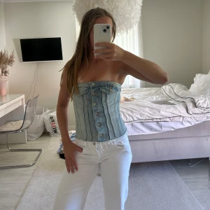 Jeans korsett-topp med knappar - Säljer en snygg axelbandslös korsett-topp i ljusblå denim med knappar framtill. Toppen har markerade sömmar och rå kant nertill för en cool vibe. Perfekt att styla med höga byxor eller kjol för en trendig look.
