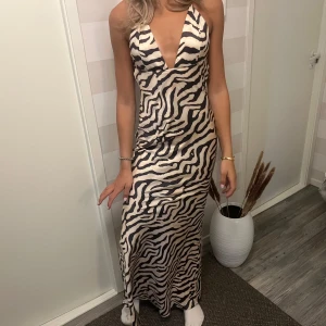 Zebramönstrad långklänning med öppen rygg - Supersnygg långklänning med zebramönster i svart och beige. Klänningen har djup v-ringning, tunna axelband och öppen rygg med knyt. Materialet är glansigt och känns lyxigt, perfekt för dig som vill sticka ut. Sitter figurnära och ger en riktigt cool look.