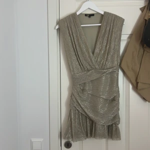 Glittrig kortklänning från Maje - Supersnygg glittrig kortklänning från Maje i champagnefärgad ton. Djup v-ringning, omlott-detalj och plisserat tyg som ger en lyxig känsla. Klänningen har dragkedja i sidan och volang nertill för extra flärd. Perfekt för dig som vill vara extra piffig till fest😍 denna klänning är köpt på NK i Stockholm för 3890kr🤍 jag har endast använt klänningen vid ett tillfälle. Den är som ny🤩