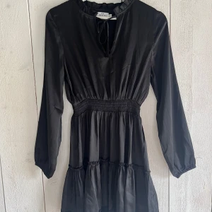 Svart klänning från NA-KD - Supersnygg svart kortklänning från Iva Nikolinas kollektion med NA-KD. Klänningen har långa ärmar, markerad midja och volangdetaljer nertill. Den har v-ringning med knytband. Köparen står för frakt💕