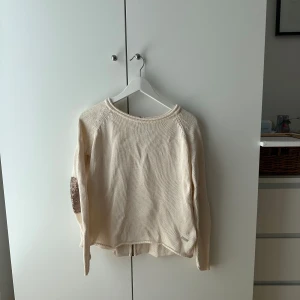 Beige stickad kofta från Monrow - Beige stickad kofta från Monrow med lång ärm och coola bruna armbågslappar. Koftan har dragkedja bak och en avslappnad passform. Perfekt för lager-på-lager och enkel att styla till olika looks.