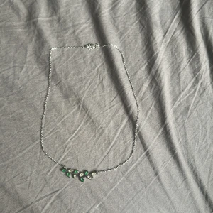  halsband med gröna och silverfärgade blad - Snyggt halsband  med tunn silverfärgad kedja och ett hänge format som en gren med små blad i grönt och vitt. Perfekt för dig som gillar naturinspirerade smycken och vill ha något unikt som sticker ut. Den är även allergivänlig. 