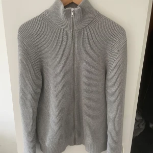 Grå stickad kofta från East West - Snygg grå stickad kofta från East West i storlek S. Modellen har hög krage och hel dragkedja framtill. Ribbstickad struktur ger en clean och stilren look, perfekt för lager-på-lager. Passar dig som gillar enkel och tidlös stil. ABS! Pris går att diskutera. Koftan är använd cirka 8 gånger. Passar as bra till vilken färg som helst på jeans och andra sorters byxor. 