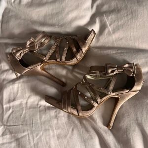 Champagnefärgade pumps med rosett och strass - Champagnefärgade pumps med hög klack, öppen tå och smala remmar dekorerade med strass. Snygg rosettdetalj på sidan och dragkedja bak. Skorna är i satinliknande material och perfekta för att lyfta din outfit.