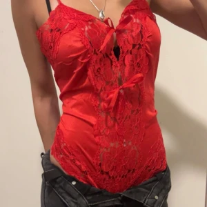 Röd spetsig bodysuit  med rosetter - Snygg röd bodysuit med spetsdetaljer och två knytband framtill. Bodysuiten har blommönstrad spets längs hela framsidan och tunna axelband. Materialet är glansigt och känns lyxigt.