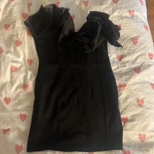Svart kort klänning med volang H&M - Supersnygg svart kort klänning från H&M med en axel och stora volangdetaljer upptill. Tight passform som framhäver figuren och ger en cool look. Perfekt för dig som vill sticka ut med en unik och trendig stil.