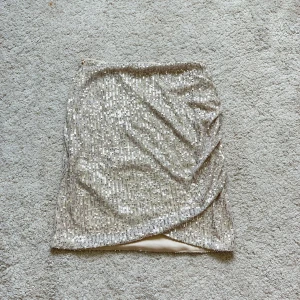 Kjol med paljetter - Snygg kjol som tyvärr inte har kommit till användning. Den är champagnefärgad, har paljetter och en omlottdesign på framsidan. Går även att använda som bandeau-topp! Kjolen är i toppenskick. Skriv om ni har frågor!