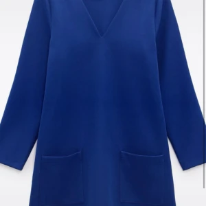 Blå klänning med v-ringning - En stilren blå klänning med v-ringad hals och långa ärmar. Modellen är rak och har två stora fickor framtill. Perfekt för dig som gillar enkel och modern design.