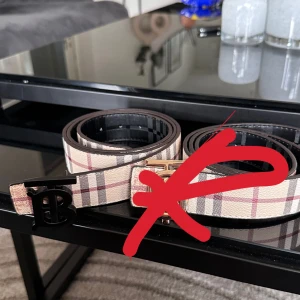 Burberry rutiga bälten med TB-spänne - Två snygga Burberry-bälten med klassiskt rutigt mönster. Ena bältet är beige med röda och svarta rutor, det andra helsvart med diskret rutmönster. Båda har ikoniskt TB-spänne i svart eller guld. Tillverkade i skinn och perfekta för att lyfta din outfit.