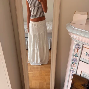 Vit långkjol med volang - Säljer en superfin vit långkjol med bred resår i midjan och volangdetaljer. Kjolen har en luftig och flowy känsla, perfekt för varma dagar. Materialet är lätt och ger en avslappnad vibe. Passar perfekt till crop tops eller stickade tröjor.