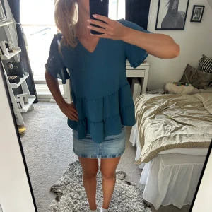 Blå volangblus med v-ringning - Säljer en blå blus med v-ringad hals och volangdetaljer i flera lager. Blusen har korta, luftiga ärmar och en loose passform som ger en avslappnad vibe. Perfekt att styla med jeans eller kjol för en trendig look.