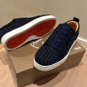 Mörkblå Christian Louboutin sneakers - Snygga mörkblå sneakers från Christian Louboutin i mocka med ikoniska röda sulor. Skorna har snörning och coola nitar på tån för en edgy vibe. Vit platt sula och lyxig känsla, perfekt för dig som vill sticka ut med exklusiva detaljer.