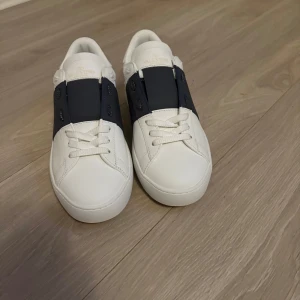 Valentino Open sneakers vit/marinblå - Snygga Valentino Open sneakers i vitt skinn med bred marinblå rem över vristen. Klassisk rund tå, vita snören och diskreta nitar på hälen. Lyxig känsla och stilren design, perfekt för dig som vill sticka ut med exklusiva detaljer. Helt nya, endast testade, säljer då de var för små.