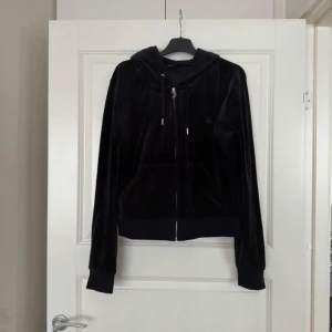 Svart velourjacka Juicy Couture XL - Svart velourjacka från Juicy Couture i storlek XL. Klassisk hoodie med dragkedja, huva med snörning och kängurufickor framtill. Mjuk och glansig yta som ger en lyxig känsla, perfekt för en chill och trendig look. Fint skick men lite nopprig i mudden se bild! Går dock bort med en nopper maskin! 