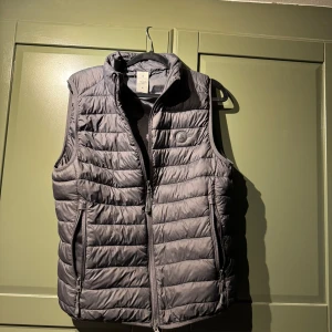 Grå dunväst från Scalpers, M - Snygg grå dunväst från Scalpers med quiltad design och hög krage. Västen har två dragkedjeförsedda fickor och en diskret logga på bröstet. Perfekt att slänga över en hoodie eller tröja när det är kyligt ute.