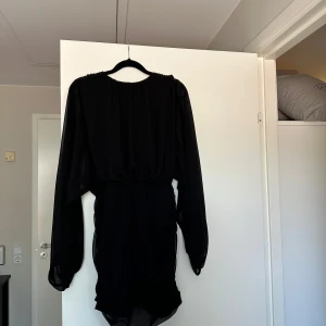 Svart kort klänning från NA-KD - Snygg svart kort klänning från NA-KD med långa, transparenta ärmar och rynkade detaljer längs kjolen. Klänningen har rund halsringning och dragkedja i ryggen. Perfekt för dig som vill ha en stilren och trendig look.