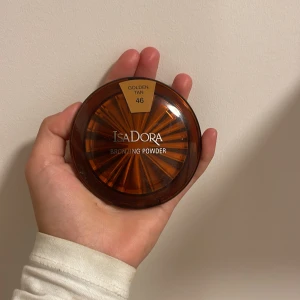IsaDora Bronzing Powder Golden Tan - IsaDora Bronzing Powder i nyansen Golden Tan 46. Kommer i en rund, brun plastförpackning med solstråle-design på locket. Puderfärgen är varm, gyllene brun och ger en solkysst look till ansiktet. Perfekt för att framhäva kindben och ge glow.