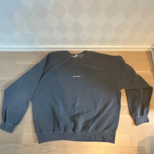 Mörkgrå sweatshirt med broderad text - En oversized mörkgrå sweatshirt med liten vit broderad text på bröstet. Tröjan har rund halsringning och långa ärmar med breda muddar. Perfekt för en chill och avslappnad stil.