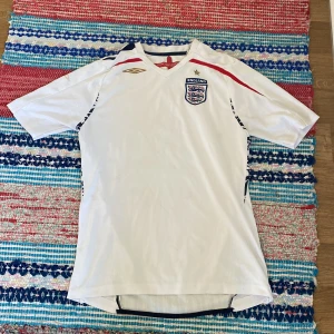 England fotbollströja Umbro 2007-2009 - England fotbollströja från Umbro, officiell landslagströja 2007-2009. Vit med röda och marinblå detaljer, broderat England-emblem och guldstjärna på bröstet. Tillverkad i ventilerande polyester med korta ärmar och sportig passform.