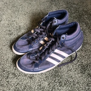 Blå Adidas sneakers med vita ränder - Snygga blå sneakers från Adidas med klassiska vita ränder på sidorna. Skorna har högt skaft, snörning och en vit sula med blå undersida. Ovandelen är i textil och syntet, perfekt för en sportig och avslappnad stil.