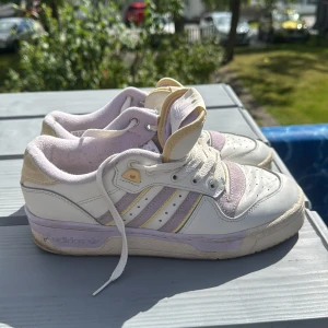 Adidas sneakers i vitt och lila - Snygga Adidas sneakers i vitt och lila med klassiska tre ränder i lila och gul detalj. Skorna har snörning, rund tå och platt sula. Ovandelen är i skinn och mocka, med Adidas-logga på tungan och sidan. Perfekta för dig som gillar retrostil och streetwear.