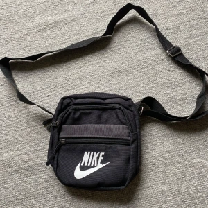 Svart Nike axelväska - Säljer en svart axelväska från nike som aldrig är använd. Väskan har en stilren design med vit Nike-logga framtill och är tillverkad i slitstark polyester. Perfekt storlek för mobil, plånbok och andra småprylar. 