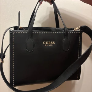 Svart handväska från Guess - Snygg svart handväska från Guess med guldiga detaljer och logga framtill. Väskan har både handtag och justerbar axelrem, dekorativa kontrastsömmar och en praktisk dragkedjeficka på baksidan. Perfekt för att bära dina essentials med stil.