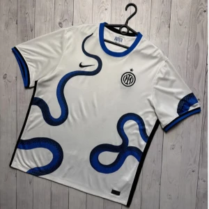 Inter Milan vit bortatröja Nike - Inter Milan bortatröja i vitt med blå och svarta ormliknande detaljer. Tröjan har korta ärmar med blå och svarta muddar, rund halsringning och klubbmärke på bröstet. Tillverkad i lätt och ventilerande material, perfekt för match eller träning.