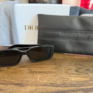 Svarta solglasögon från Christian Dior - Snygga svarta solglasögon från Christian Dior med rektangulära bågar och tydlig logga på sidorna. Glasögonen har en chunky design och är tillverkade i blank plast. Perfekta för dig som vill ha en stilren och trendig accessoar.