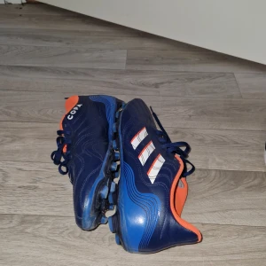 Adidas Copa blå fotbollsskor - Säljer ett par Adidas Copa fotbollsskor i mörkblått med orange foder och vita ränder på sidan. Skorna har snörning och är tillverkade i skinn med klassisk rund tå och dobbar för gräsplan. Perfekta för dig som vill sticka ut på planen!