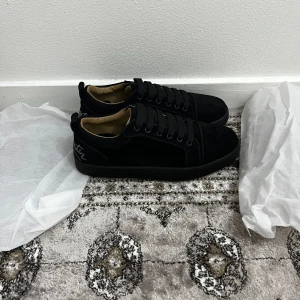 Louboutin skor - Snygga svarta sneakers från Louboutin i mjuk mocka med klassisk röd sula och broderad logga på hälen. Skorna har rund tå, platt sula och svarta snören. Perfekta för dig som vill ha en stilren och lyxig look med streetkänsla.