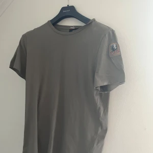 Grå t-shirt från Parajumpers - Snygg grå t-shirt från Parajumpers med klassisk rund hals och korta ärmar. På vänster ärm sitter ett broderat Parajumpers-märke som ger en cool detalj. T-shirten är i mjuk bomull och passar perfekt till jeans eller shorts. Orginal pris 1 200 kr Storlek S men passar också M