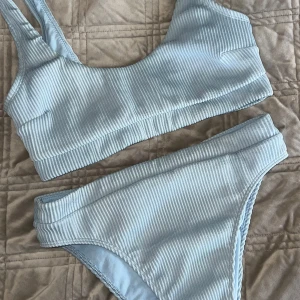 Ljusblå ribbad bikini - Superfin ljusblå bikini med ribbad struktur. Toppen har breda axelband och sportig form, medan bikinitrosan har hög midja och breda sidor. Perfekt för strandhäng eller poolparty! Materialet känns mjukt och stretchigt.
