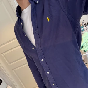 Mörkblå Polo Ralph Lauren Skjorta - Snygg blå skjorta från Polo Ralph Lauren i slim fit-modell, storlek M. Skjortan är i 100% bomull och har klassisk button down-krage, vita knappar och den ikoniska gula loggan broderad på bröstet. Perfekt nu för sommaren. Riktigt härlig färg!