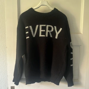 Svart stickad tröja med text - Svart stickad tröja med vit text som säger 'EVERY DAY HAPPY' över bröstet och ärmarna. Tröjan har rund halsringning, ribbade muddar och en loose fit. Perfekt för dig som gillar statement-plagg och vill ha en chill vibe.