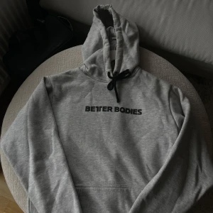 Grå hoodie från Better Bodies XS - Snygg grå hoodie från Better Bodies i storlek XS. Tröjan har svart snörning i huvan och svart tryck på bröstet. Perfekt för chill dagar eller till gymmet. Mjuk och skön med klassisk känguruficka framtill.