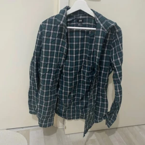 Rutig grön skjorta från H&M - Snygg rutig skjorta från H&M i grönt, svart och vitt med diskreta röda linjer. Skjortan har klassisk krage, knäppning framtill och långa ärmar. Perfekt för en chill och avslappnad stil. Tillverkad i mjuk bomull som känns skön mot huden.