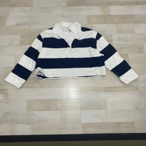 Randig croppad pikétröja i blått och vitt - Snygg croppad pikétröja med breda marinblå och vita ränder. Tröjan har klassisk krage, knappar vid halsen och långa ärmar. Mjuk insida och relaxed fit, perfekt för en sportig och chill look. I dem två sista bilderna kan ni se att det finns några fläckar.