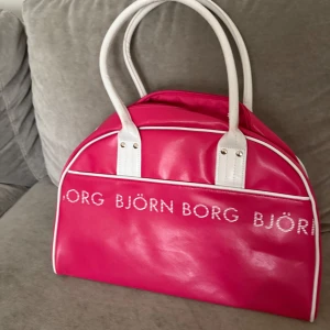Rosa handväska från Björn Borg - Cool rosa handväska från Björn Borg med vita detaljer och handtag. Väskan har ett stilrent tryck med loggan framtill och dragkedja upptill. Tillverkad i syntetmaterial och har ett rymligt fack med innerficka. Perfekt för dig som vill sticka ut med färg.